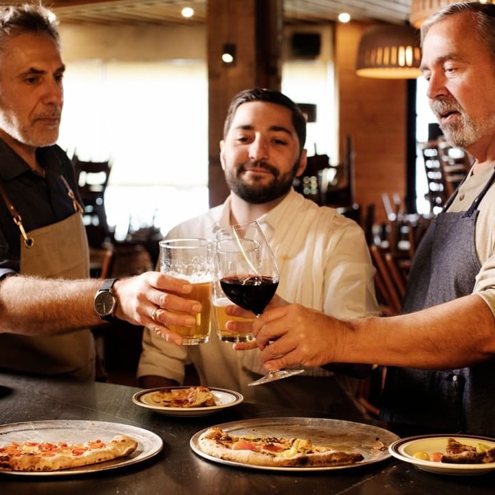 Three men toast with their beverages over a counter covered with wood-fired pizzas